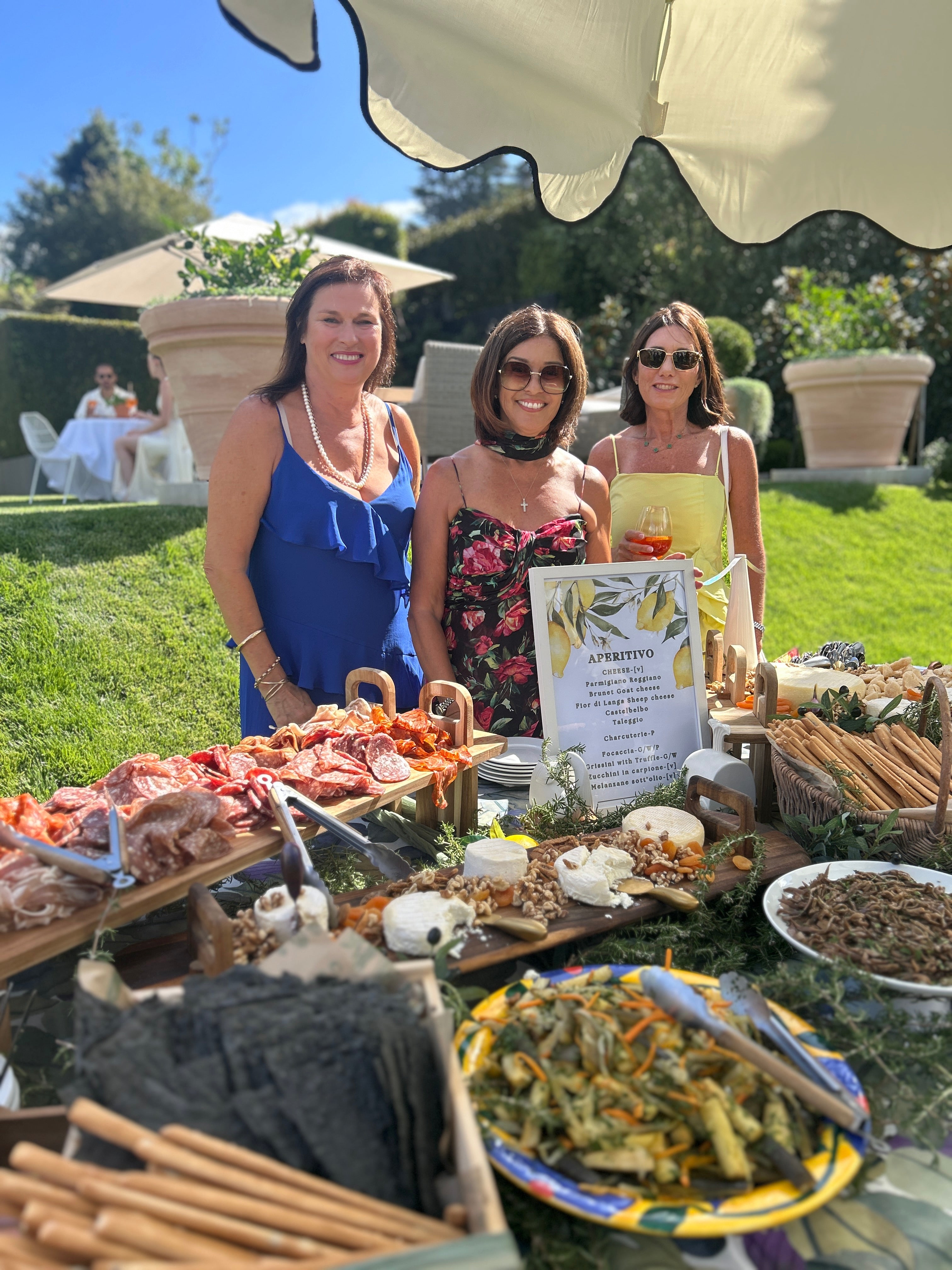 Three women standing behind a table with food and drinks outdoors