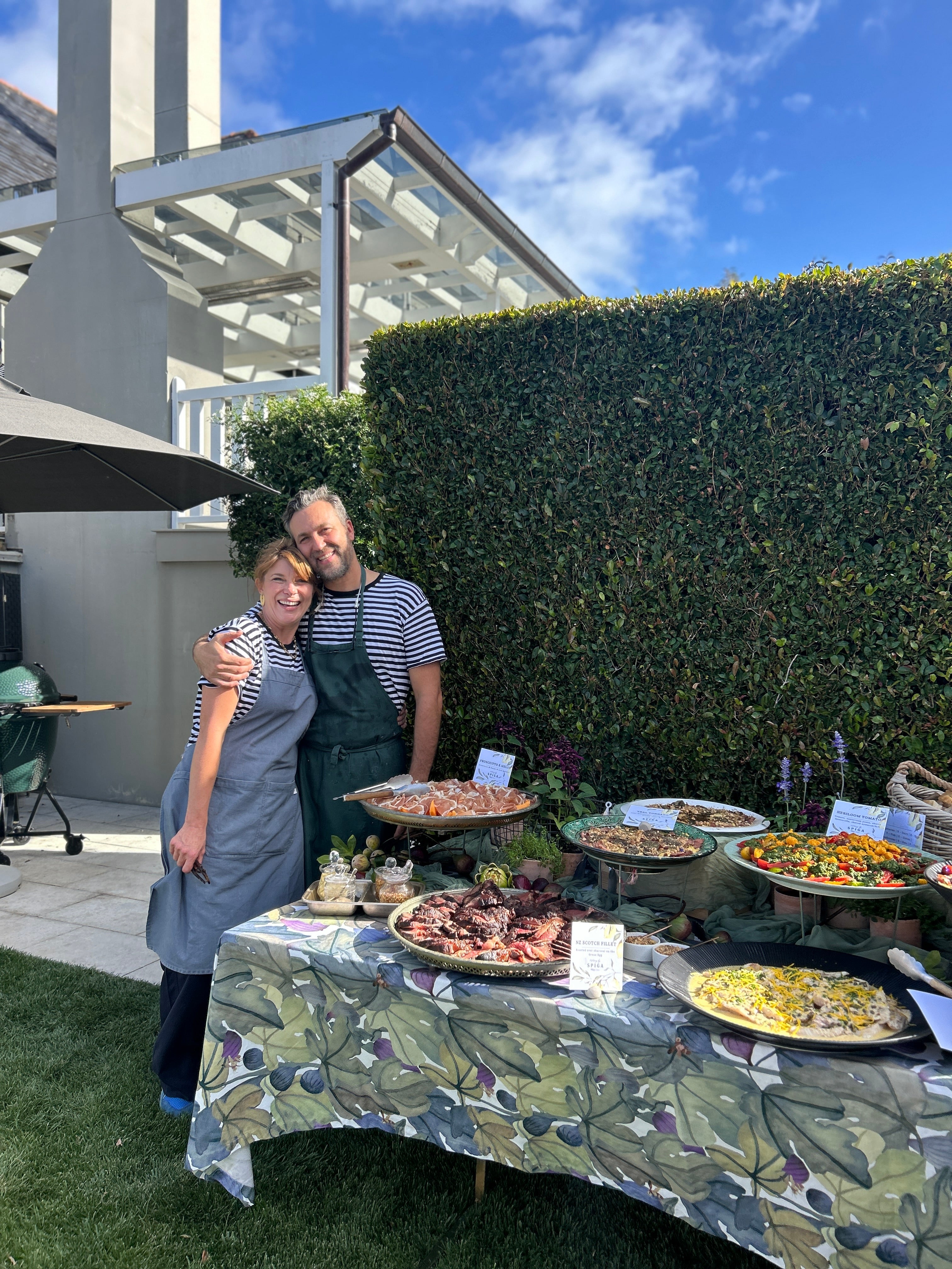 Two people standing behind a table with food outdoors on a sunny day.