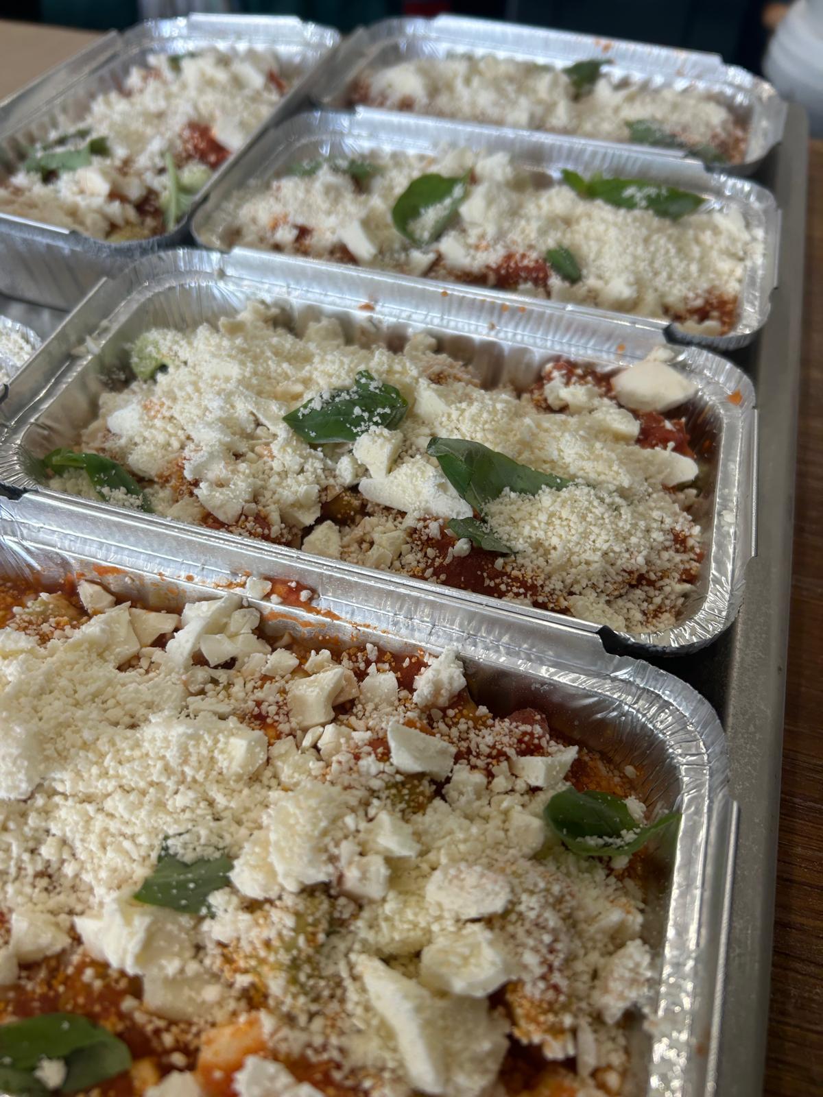 Aluminum trays with pasta dishes topped with cheese and basil leaves on a wooden surface.