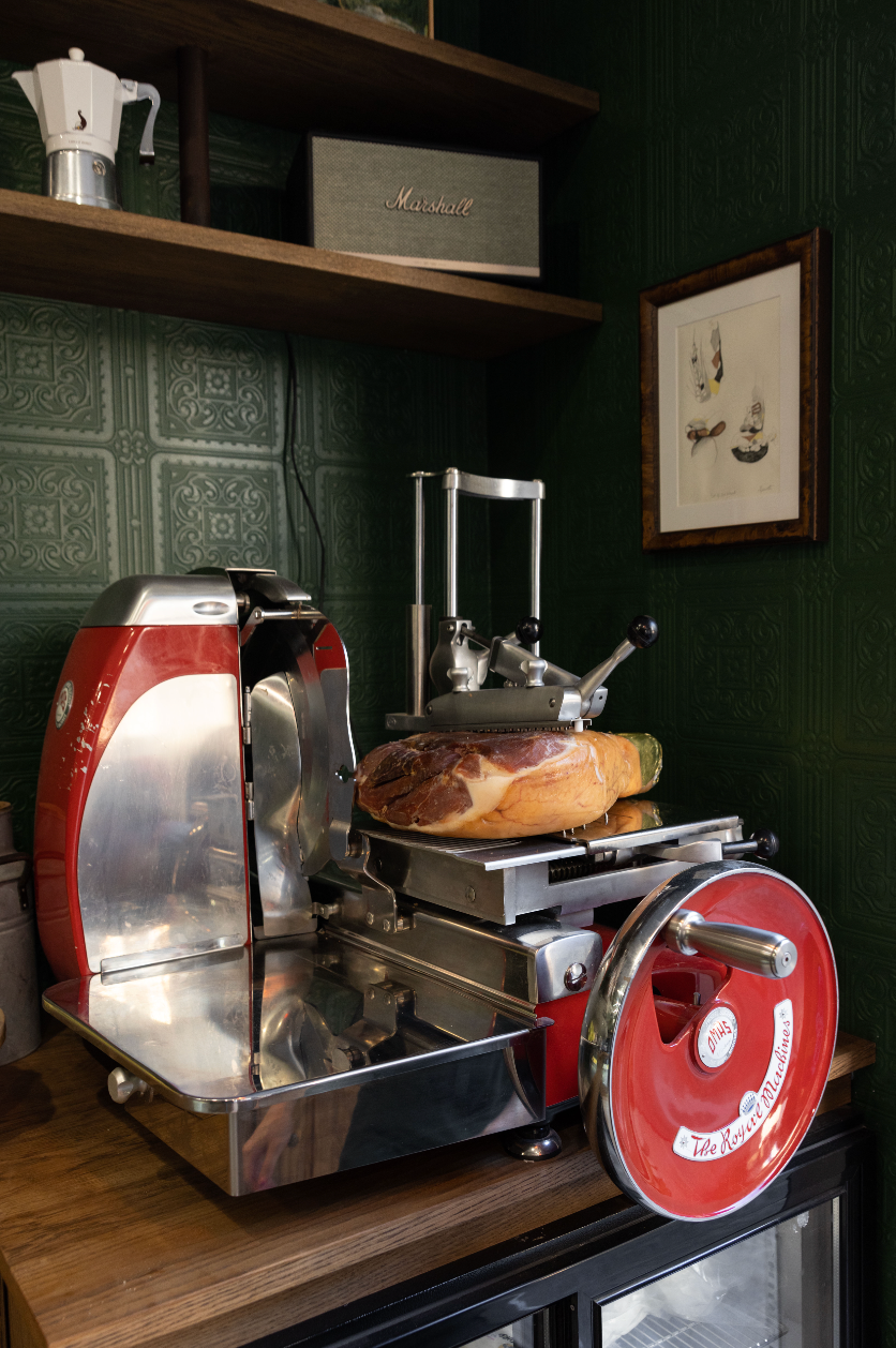 Sliced meat on a meat slicer with a dark green wall and shelf in the background